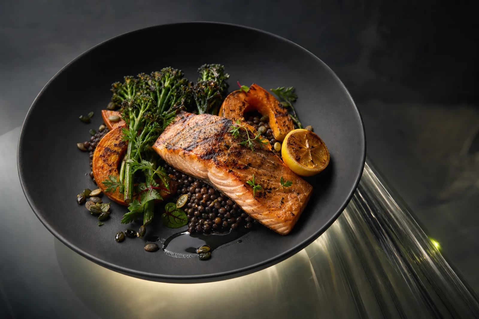 A grilled salmon fillet with black lentils, charred broccolini and roast squash in a dark bowl